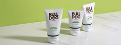 Three Bulldog Skincare products—a face wash, moisturizer, and post-shave balm—are displayed on a white marble surface with a green background.