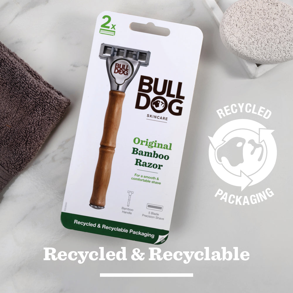 An Original Bamboo Razor is packaged in a white box labeled with a recycled icon and Recycled & Recyclable Packaging, displayed on marble beside skincare essentials: a towel and pumice stone.