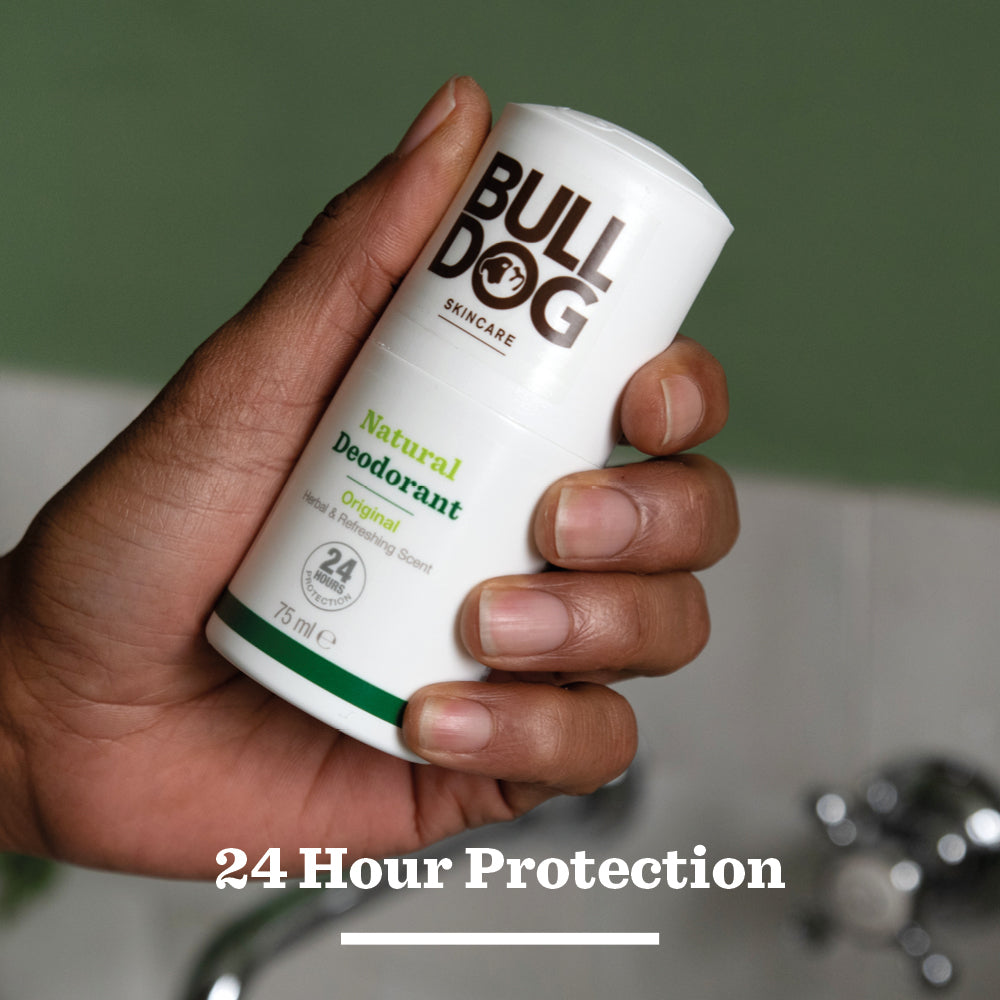 A hand holds a stick of Original Natural Deodorant from Bulldog Skincare—one of the top mens grooming picks—against a blurred green and white background. Text below reads: 24 Hour Protection.