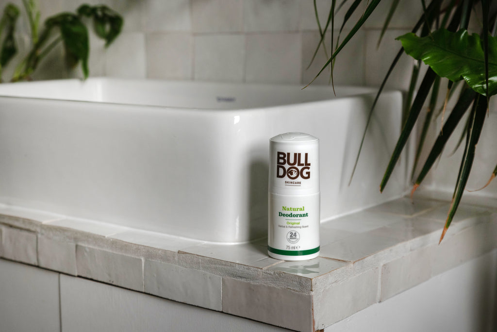 A stick of Original Natural Deodorant, offering 24-hour odour protection, rests on the edge of a white bathroom sink surrounded by green leafy plants.