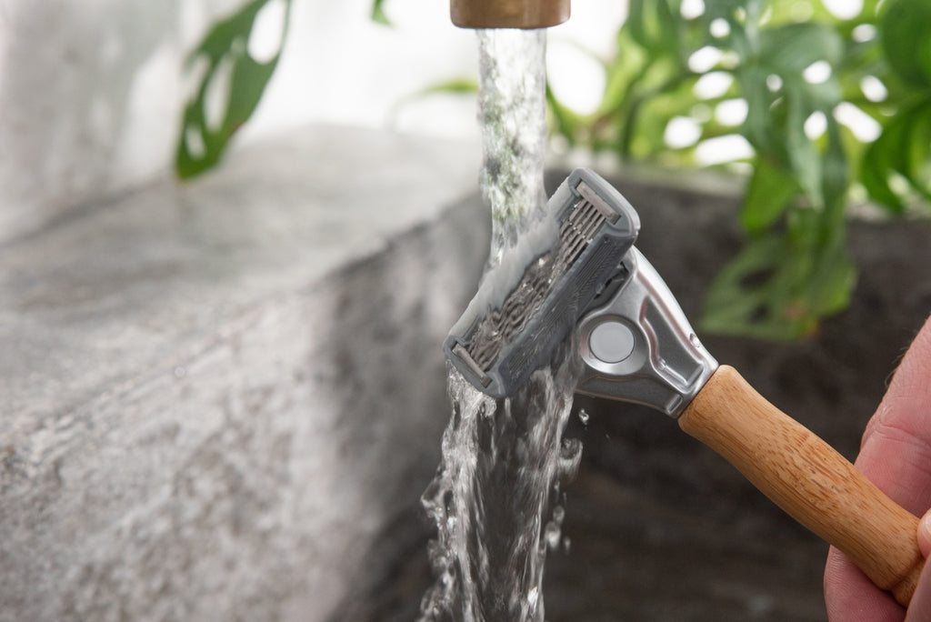 A close-up of a hand rinsing the Original Bamboo Razor with tempered steel blades under running water from a faucet, with green plants blurred in the background.