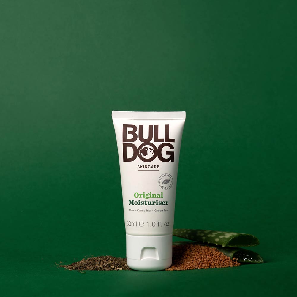 A tube of Travel Original Moisturiser stands upright on a green background, surrounded by aloe vera, seeds, and green tea leaves, emphasizing its natural ingredients. The white tube features black and green text.