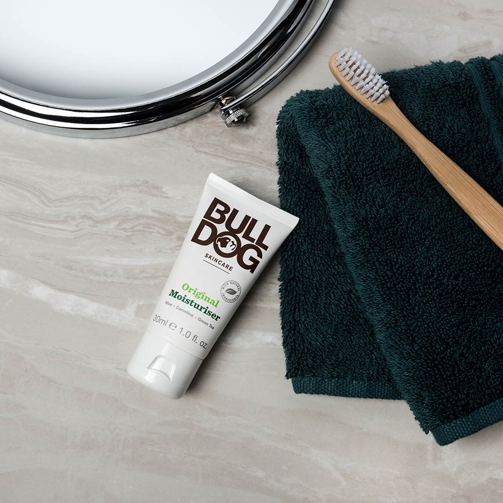 A Travel Original Moisturiser tube made for men’s skincare rests on a dark green towel beside a wooden toothbrush, all set atop a beige marble countertop near a round mirror.