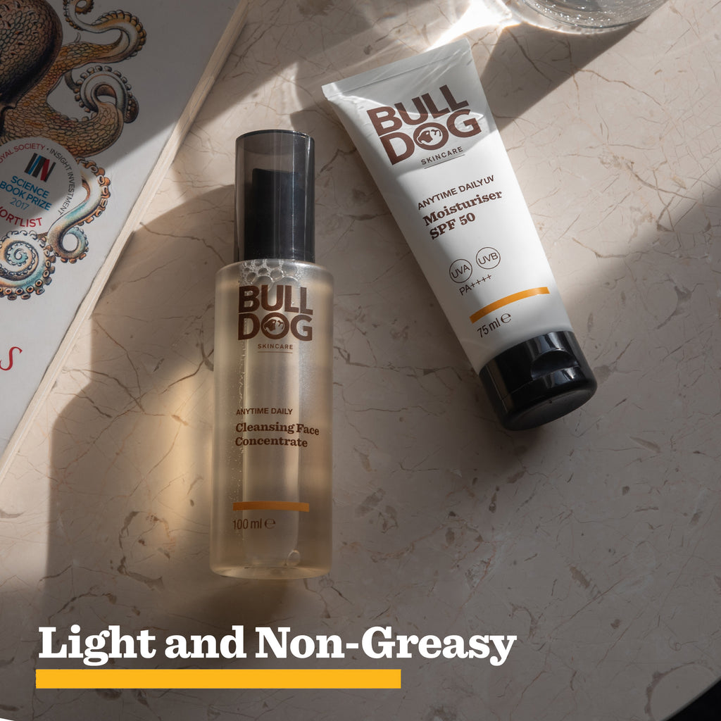 The Bulldog Skincare Anytime Daily UV Moisturiser SPF 50 tube sits on a sunlit surface beside a partially visible magazine. Text below reads: Light and Non-Greasy for all-day hydration.