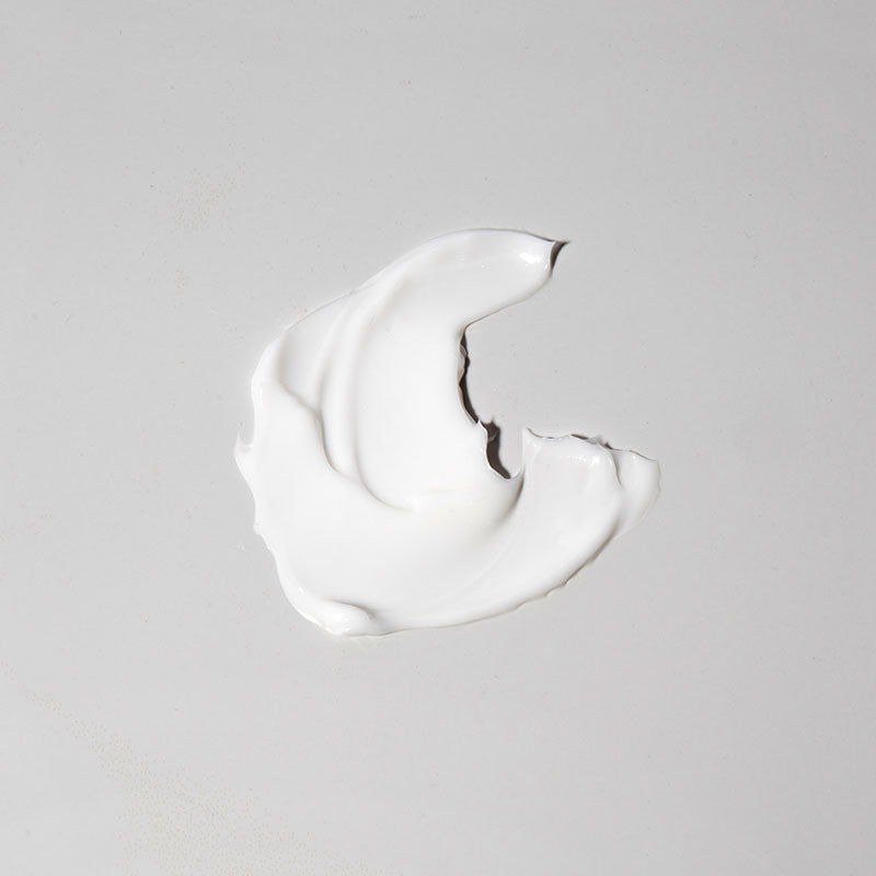 A smear of Travel Original Moisturiser, crafted with natural ingredients, is spread on a plain light gray surface in a crescent or abstract shape with smooth, slightly textured edges.